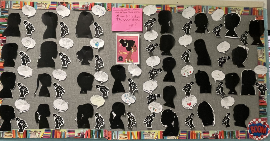 Bulletin board with 25 children's silhouettes on black construction papers, holding microphones. And a color photo of Nina Simone and question that asks, "Inspired by Christian Robinson and the great Nina Simome, What lyric does the wold need to hear?" Speeck bubble responses around the silhoette read things like "go peace and love". 